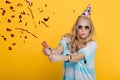 Portrait of funny blond woman in birthday hat and red confetti on yellow background. Celebration and party. Royalty Free Stock Photo