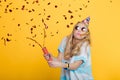 Portrait of funny blond woman in birthday hat and red confetti on yellow background. Celebration and party. Royalty Free Stock Photo