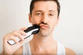 Portrait of funny brutal young man shaving his mustache with modern electric razor and looking at camera indoors. Royalty Free Stock Photo