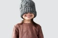 Portrait of funny cheerful little girl hidden the eyes in winter warm gray hat, joyful smiling and wearing sweater isolated on a Royalty Free Stock Photo