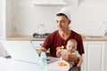 Portrait of funny concentrated handsome freelancer male wearing burgundy t shirt, posing in white kitchen, sitting in front of