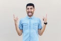 Portrait of funny crazy handsome young bearded man in blue shirt standing with rock sign gesture, tongue out and closed eyes Royalty Free Stock Photo