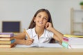 Portrait of funny and cute schoolgirl with dreamy expression sitting at desk between books.