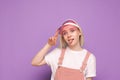 Portrait of a funny girl in a pretty casual dress, wearing a pink cap and making a funny face on a blue background. Funny teen Royalty Free Stock Photo