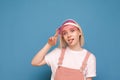 Portrait of a funny girl in a pretty casual dress, wearing a pink cap and making a funny face on a blue background. Funny teen Royalty Free Stock Photo