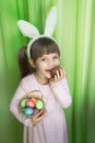 A portrait funny little girl with brown hair and a toothless smile, wearing bunny ears, eating chocolate egg and holding Royalty Free Stock Photo