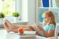 Portrait of funny little school girl relaxing reads book at the table in room at home