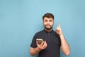 Portrait of a funny man with a beard and a phone in his hand on a blue background, looking into the camera and making a funny face Royalty Free Stock Photo