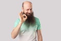 Portrait of funny middle aged bald man with long beard in light green t-shirt standing, winking and looking at camera with Ok sign Royalty Free Stock Photo