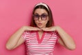 Portrait of funny optimistic woman wearing striped T-shirt and sunglasses posing isolated over pink background, standing with pout