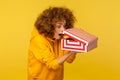 Portrait of funny, surprised, curious curly-haired woman in hoodie looking into gift box, unwrapping present