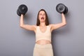 Portrait of funny weak woman wearing top and leggins holding heavy barbells in hands raised arms, screaming, having hard training