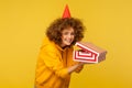 Portrait of funny woman with party cone hat looking at camera with cunning smile and holding unpacked gift box Royalty Free Stock Photo