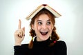 Portrait of funny young smiling student girl with an open book on her head holding her point finger up having an idea. Reading and