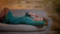 Portrait of ginger curly-haired caucasian girl lying on sofa and talking on smartphone gladly on cosy home background.