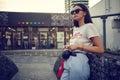 Portrait of a girl in dark sunglasses posing near a city mall. Dressed in white t-shirt, blue trousers, black waist bag Royalty Free Stock Photo