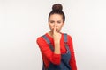 Portrait of girl with hair bun in overalls pointing nose doing lie gesture, suspecting trickster in falsehood, disbelief