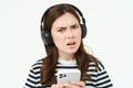 Portrait of girl in headphones with smartphone, looks confused at camera, frowns with perplexed face, stands over white Royalty Free Stock Photo