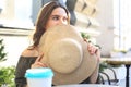 Portrait of a girl hiding her face behind a straw hat while sitting in summer street cafe