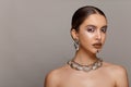 Frontal beauty portrait of a girl with makeup and jewelry applied on face, with bare shoulders, over gray background.