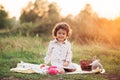 Portrait of a girl on a meadow in the sunset sunlight. Little girl in a light overalls sits on a blanket next to basket with harve