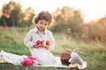 Portrait of a girl on a meadow in the sunset sunlight. Little girl in a light overalls sits on a blanket next to basket with harve