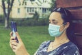 Portrait of a Girl with medical mask at park with smartphone during covid quarantine. A young girl in a medical mask sits on bench