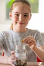 Portrait Of Girl Putting Coins Into Glass Jar Labelled Savings At Home