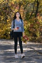 Portrait of girl student walking along the alley of the autumn park. Young beautiful woman with phone. Vertical frame