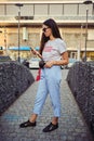 Portrait of girl in sunglasses posing near a city mall using smartphone. Dressed in white t-shirt, blue trousers, black Royalty Free Stock Photo