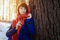 Portrait of a girl in a Sunny winter forest, which stands near a tree in a blue jacket and a red scarf