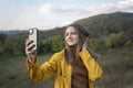 Portrait of girl taking selfie on smartphone on mountain background. Young woman in yellow park jacket enjoys beauty of autumn