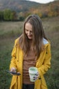 Portrait of girl with thermo cup and smartphone on autumn landscape background. Young woman drinks tea while walking in nature Royalty Free Stock Photo