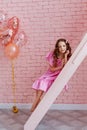 Portrait of girls with long curled hair in a pink dress on a pink background. Sitting on the stairs next to a bunch of balloons