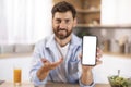 Portrait of glad adult caucasian man with beard shows phone with blank screen in kitchen interior