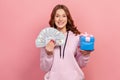 Portrait of glad brunette teenage girl in hoodie holding bunch of dollars and wrapped gift box, choice, happy winner Royalty Free Stock Photo