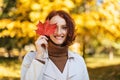Portrait of glad pretty caucasian young red-haired lady in raincoat puts red leaf to eye, enjoys freedom Royalty Free Stock Photo