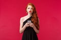 Portrait of gloomy upset gorgeous redhead girl in black dress, sobbing and grimacing unhappy, holding mobile phone Royalty Free Stock Photo