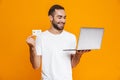 Portrait of good-looking man 30s in white t-shirt holding silver laptop and credit card, isolated over yellow background Royalty Free Stock Photo