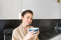 Portrait of good-looking young woman, drinking coffee in the kitchen, enjoying her morning routine and smiling at the Royalty Free Stock Photo