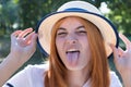 Portrait of gorgeous teenage girl in yellow hat and with red hair showing her tongue outdoors on sunny summer day