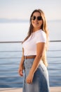 Portrait of gorgeous woman in sunglasses in front of sea. Attractive female in denim style stand on balcony in sunset