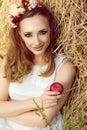 Portrait of gorgeous woman in white sundress sitting at the haystack with garland of flowers on her head, holding a red apple Royalty Free Stock Photo