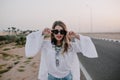 Portrait of graceful stylish girl in black sunglasses and white blouse having fun next to highway in summer. Charming