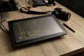 A portrait of a graphical drawing pen tablet and its stylus lying on a wooden table next to a photo camera. The tablet is