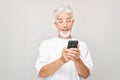 Portrait of gray-haired man in white T-shirt holding mobile phone in hand with happy smiling face. Person with smartphone Royalty Free Stock Photo