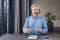 Portrait of a gray-haired senior man sitting at a desk in a home office wearing a headset and talking to the camera Royalty Free Stock Photo