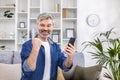 Portrait of a gray-haired senior man sitting at home on the sofa and using the phone. He looks at the camera and points Royalty Free Stock Photo