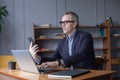 Portrait of grey hair elderly man sitting by the table with laptop and working on mobile phone Royalty Free Stock Photo