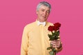 Portrait of grey haired elderly man in yellow shirt and white bow tie, holds red roses, present for his wife for birhday, poses